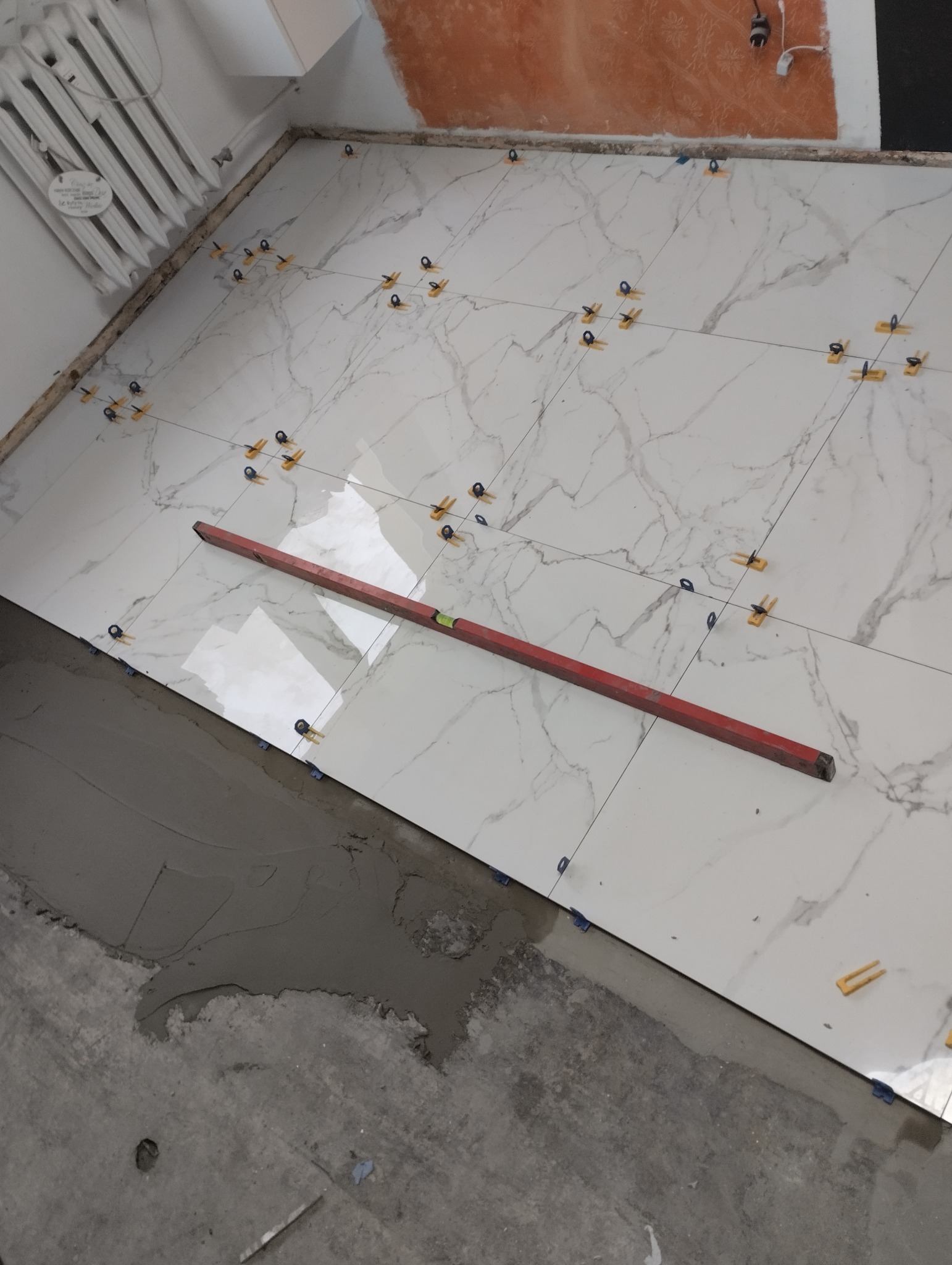 Białe płytki podłogowe z marmurkowym wzorem, układane z użyciem poziomicy i systemu poziomowania, widoczne ślady zaprawy.