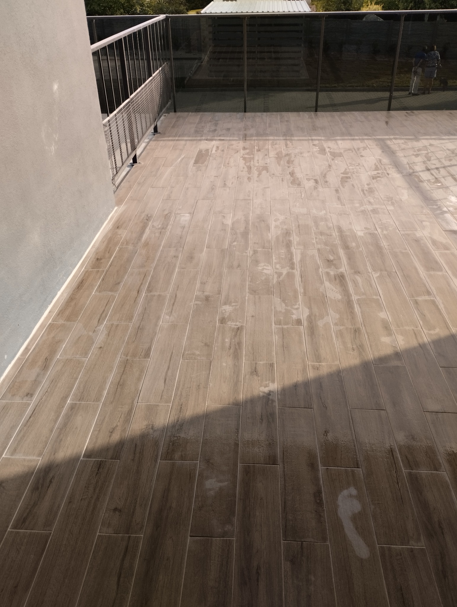 Wyłożony płytkami drewnopodobnymi balkon z metalową balustradą i fragmentem szklanej zabudowy, widoczny ślad mokrej stopy na płytkach.