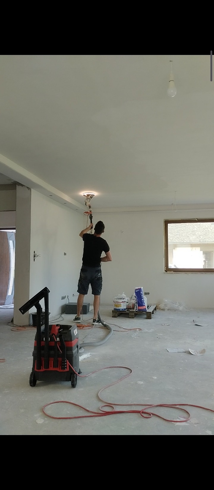 Mężczyzna szlifuje sufit szlifierką z odkurzaczem przemysłowym w pomieszczeniu w stanie surowym, widoczne puszki z gipsem i narzędzia na palecie, okno z widokiem na dach.