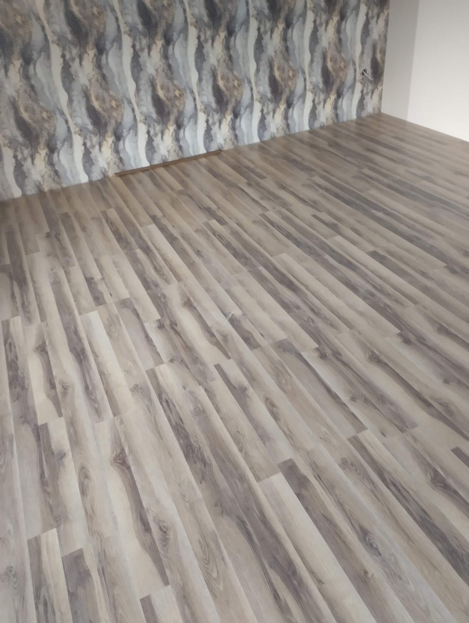 Ułożone panele podłogowe w odcieniach szarości i beżu, widoczne na tle wzorzystej tapety w podobnej kolorystyce, z fragmentem białej ściany w rogu pomieszczenia.