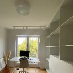 Jasny pokój biurowy z białym biurkiem, krzesłem, komputerem, wysoką zabudową regałową i dekoracyjnym wazonem z trawą pampasową, oświetlony światłem dziennym z okna.