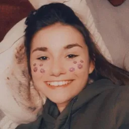 Uśmiechnięta młoda kobieta z filtrem kwiatowym na policzkach, leżąca na poduszce obok psa, ubrana w zieloną bluzę z kapturem.