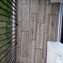Wykończony balkon z brązowymi płytkami imitującymi drewno, ułożonymi w prostokątnym wzorze, widok z góry na balustradę i fragment ściany balkonu.