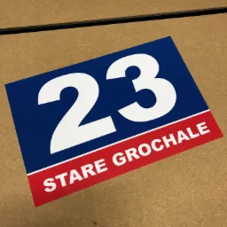 Niebiesko-czerwona tabliczka z białymi cyframi 23 i napisem STARE GROCHALE, leżąca na brązowej powierzchni.