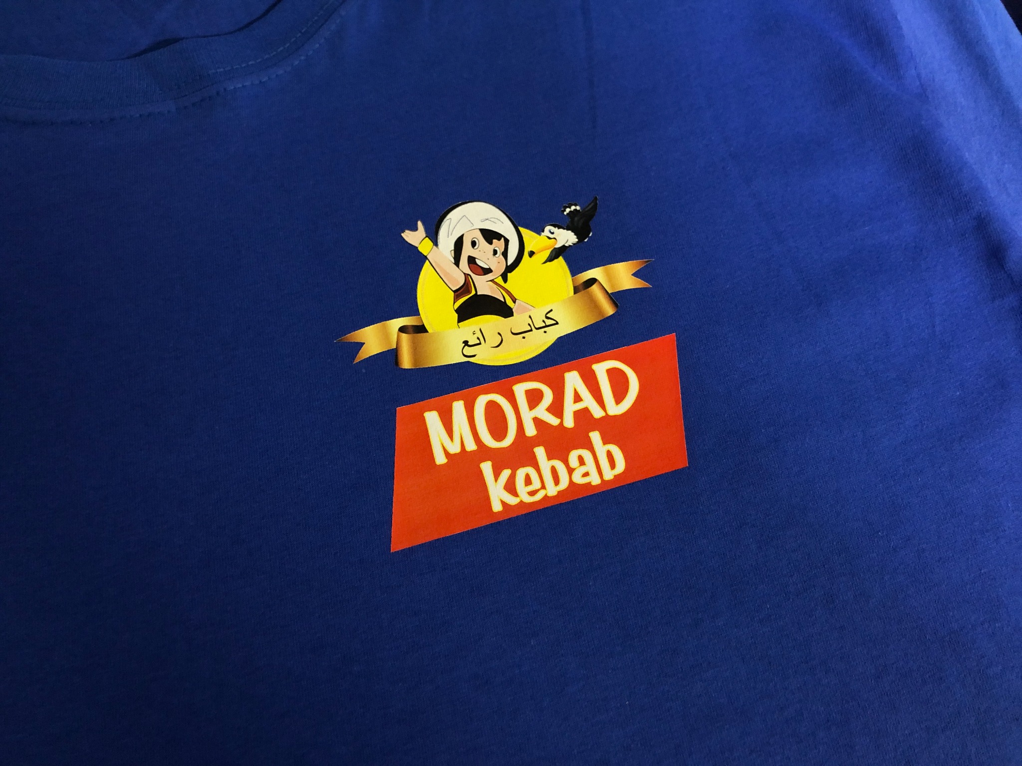 Niebieska koszulka z nadrukiem logo restauracji Morad Kebab, zawierającym postać z kreskówki, napis w języku arabskim i nazwę restauracji na pomarańczowym tle.
