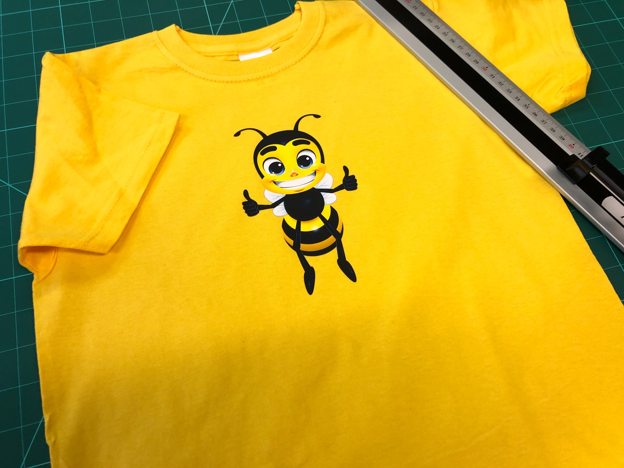 Żółta koszulka z nadrukowaną kreskówkową pszczołą z uśmiechem i kciukami w górę, leżąca na zielonej macie do cięcia obok aluminiowej linijki.