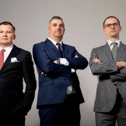 Portret trzech mężczyzn w garniturach, stojących z założonymi rękami na neutralnym tle, z różnymi wzorami i kolorami krawatów.