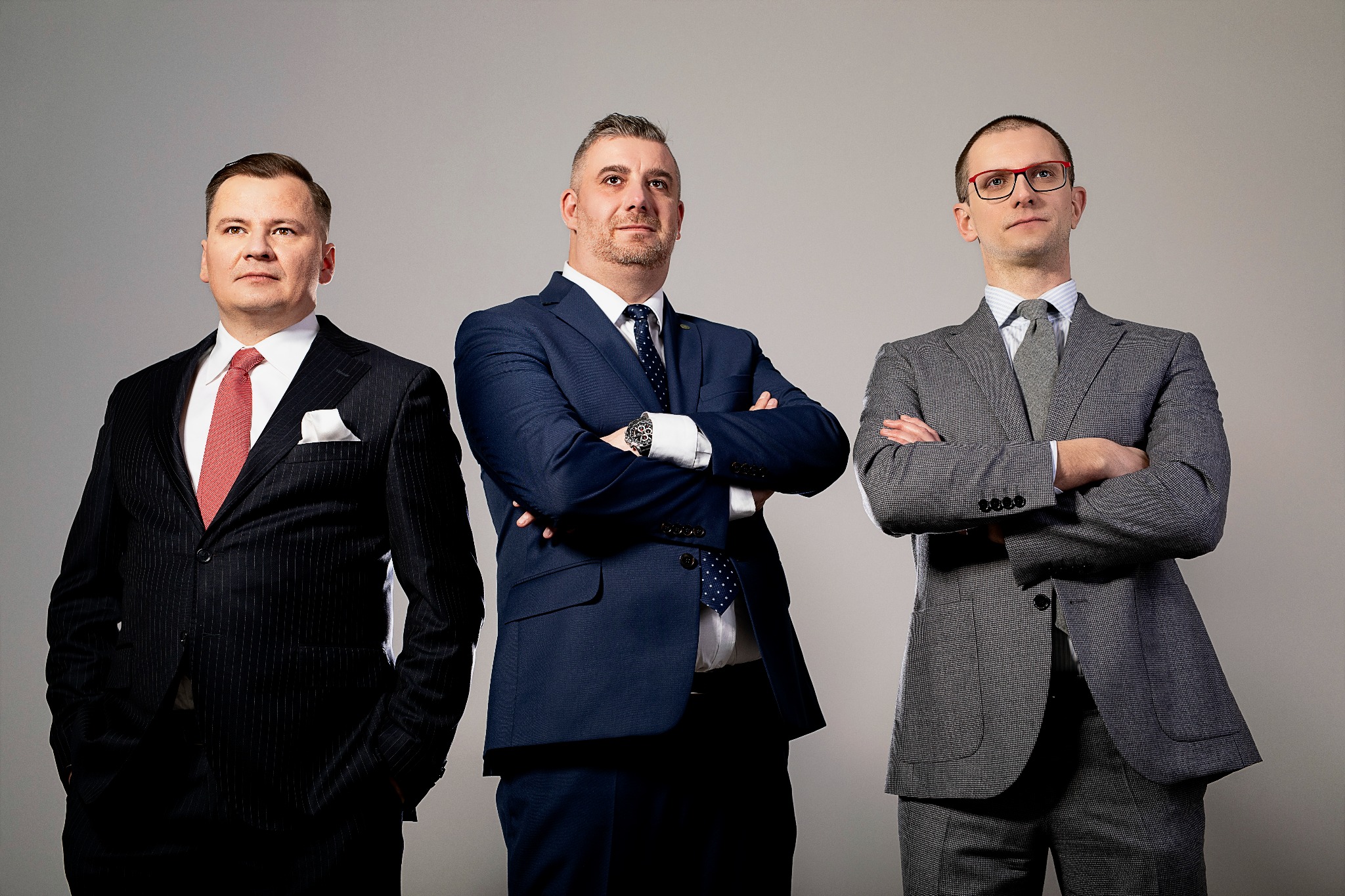 Portret trzech mężczyzn w garniturach, stojących z założonymi rękami na neutralnym tle, z różnymi wzorami i kolorami krawatów.
