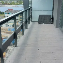 Nowoczesny balkon z szarą, płytkową podłogą, przeszkloną balustradą i widokiem na plac budowy w tle.