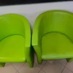 Dwa identyczne, jasnozielone fotele klubowe z ekoskóry ustawione obok siebie na kafelkach podłogowych, w tle biurowe półki z segregatorami.