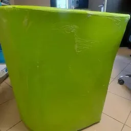 Jaskrawozielony, obity materiałem pojemnik na śmieci w biurze, z widocznymi śladami użytkowania na powierzchni, stojący obok biurka i krzesła biurowego na kółkach, w tle regał.