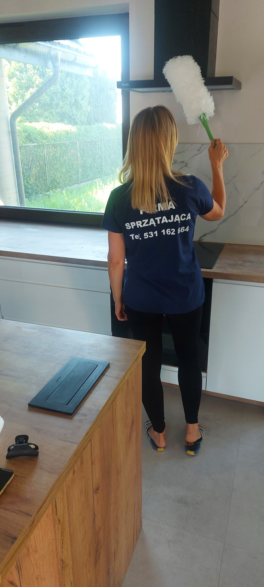 Kobieta w koszulce firmy sprzątającej czyści okap kuchenny białym, pióropuszowym odkurzaczem do kurzu w nowoczesnej kuchni z białymi szafkami i drewnianym blatem.