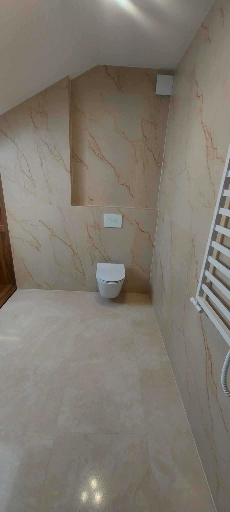 Wykończona łazienka na poddaszu z beżowymi płytkami imitującymi marmur, wisząca toaleta z podtynkową spłuczką i drabinkowy grzejnik.