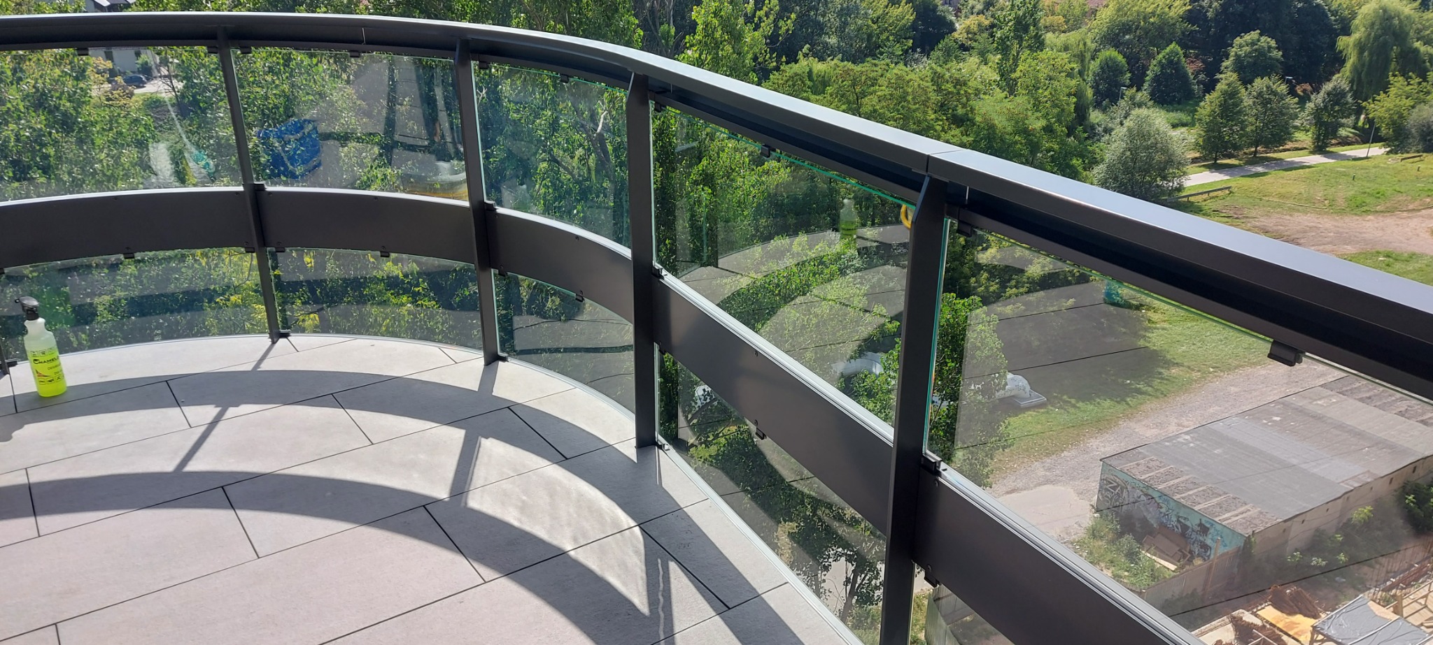 Szklana balustrada balkonu z widokiem na zieleń, na pierwszym planie butelka ze środkiem czyszczącym.