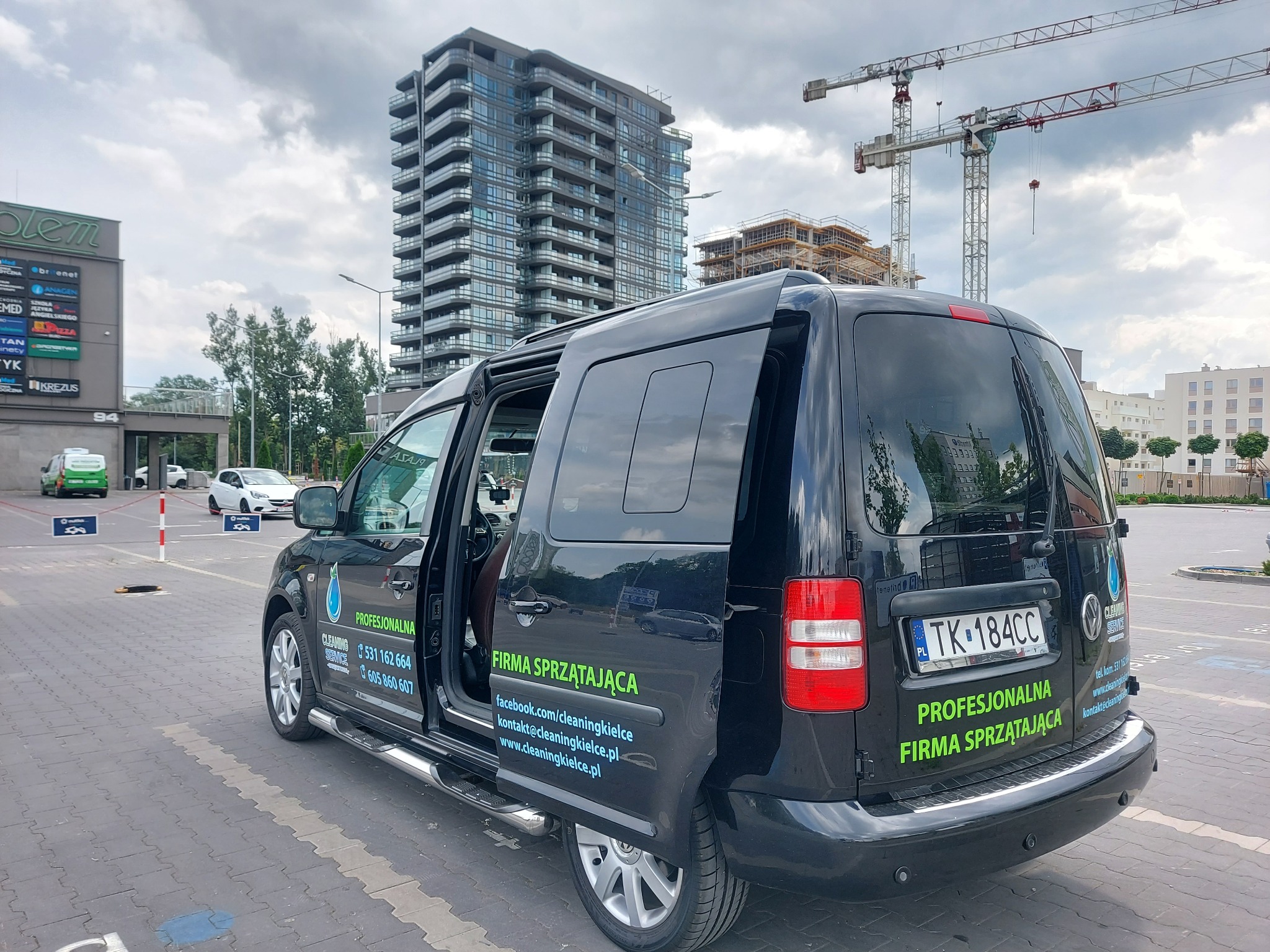 Czarny samochód dostawczy z otwartymi drzwiami, oklejony reklamą firmy oferującej profesjonalne usługi czystościowe, zaparkowany na parkingu przed budynkiem handlowym i wysokim apartamentowcem w tle.