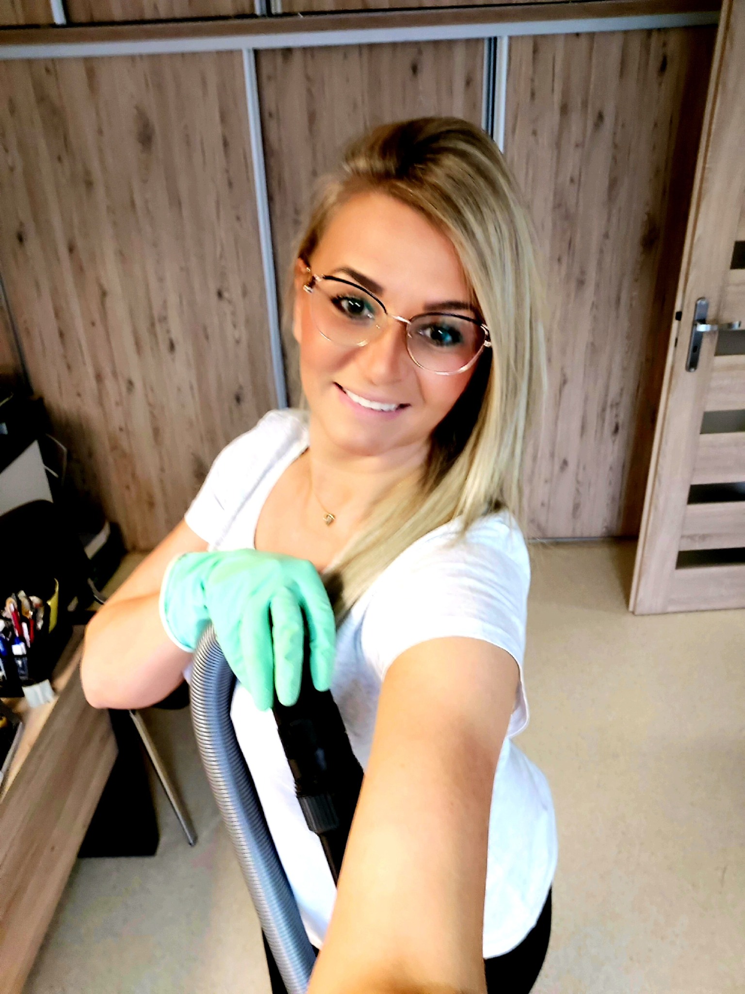 Uśmiechnięta blondynka w okularach i zielonych rękawicach trzyma rurę od odkurzacza na tle szafy przesuwnej z drewnianymi panelami.