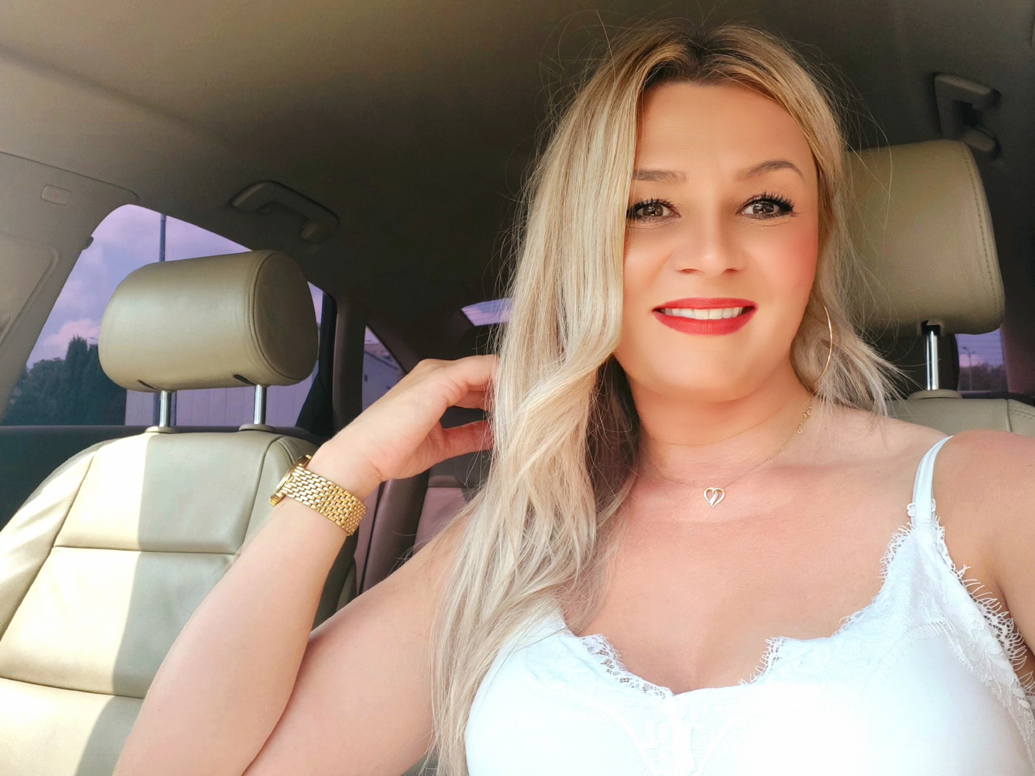 Uśmiechnięta blondynka w samochodzie, złota bransoletka i naszyjnik z serduszkiem, jasna tapicerka fotela.