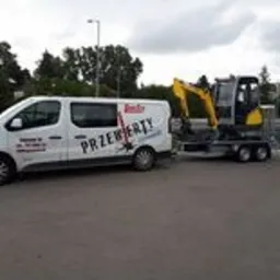 Żółta mini koparka na platformie transportowej za białym busem z napisem 'PRZEWERTY'.