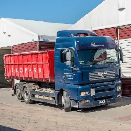 Niebieska ciężarówka MAN z czerwoną skrzynią transportową z napisem EKO PARK S.A. zaparkowana przed halą magazynową z bramą rolowaną.