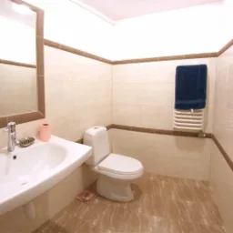 Jasna łazienka z beżowymi płytkami i brązowymi akcentami, widoczna umywalka narożna z lustrem, toaleta i grzejnik drabinkowy z niebieskim ręcznikiem.