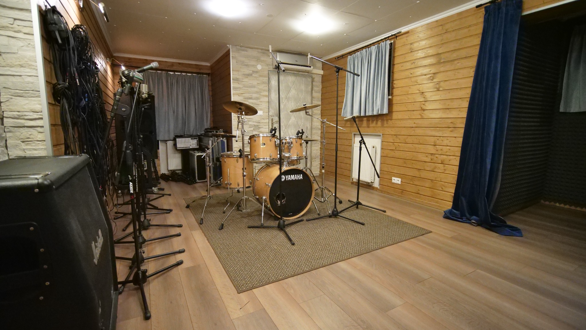 Jasne, przestronne pomieszczenie z perkusją Yamaha na dywaniku, mikrofonami na statywach, kablami i wzmacniaczem Marshall, panele akustyczne na ścianie, drewniane wykończenie ścian.