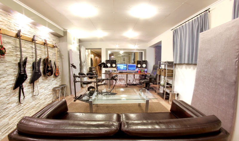 Wnętrze studia nagraniowego z brązową skórzaną sofą na pierwszym planie, na ścianie wiszą gitary elektryczne i akustyczne, w tle widoczne stanowisko pracy z dwoma monitorami i perkusją elektroniczną.