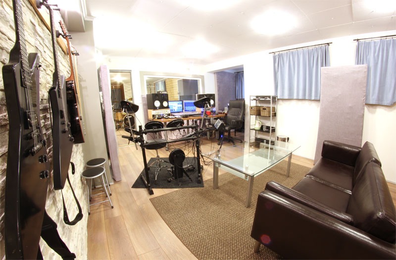 Wnętrze pokoju z instrumentami muzycznymi: gitary elektryczne na ścianie z kamienia, elektroniczna perkusja, monitory studyjne i sofa.