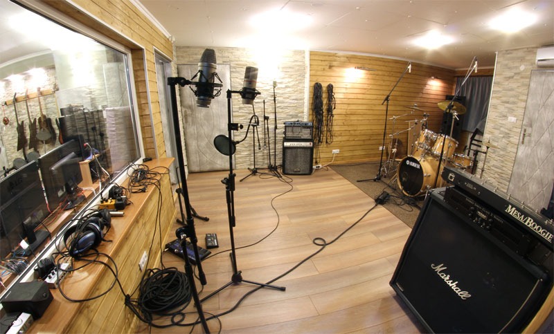 Wnętrze studia nagrań z mikrofonami pojemnościowymi, perkusją Yamaha i wzmacniaczami Marshall i Mesa Boogie, widok przez szybę na pomieszczenie z gitarami.