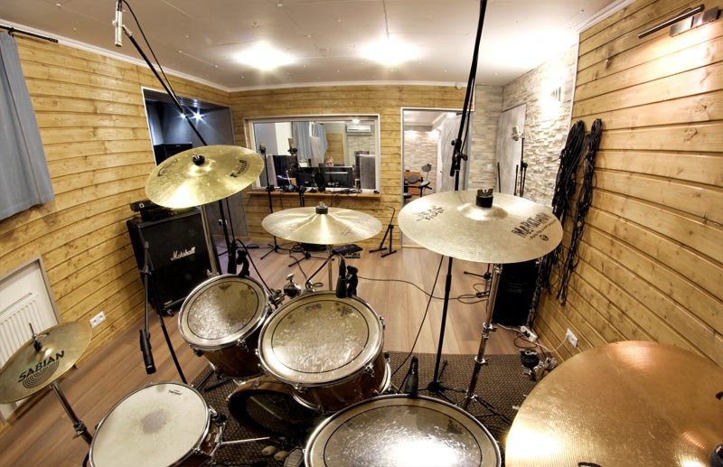 Zestaw perkusyjny z mikrofonami w wygłuszonym pomieszczeniu z drewnianymi panelami, widok z perspektywy perkusisty, przez okno widać stanowisko realizatora dźwięku.
