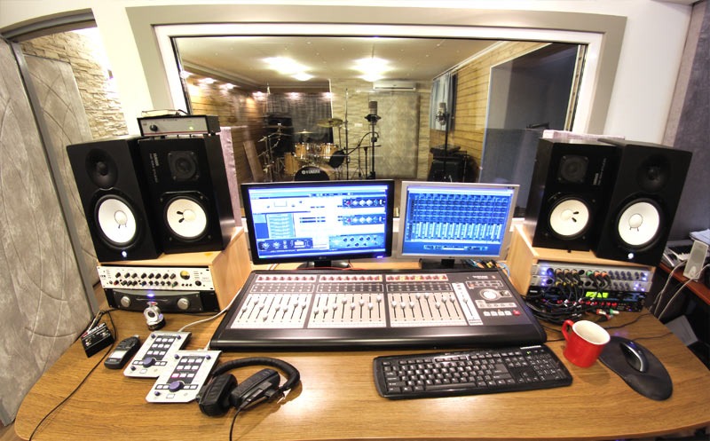 Profesjonalne stanowisko do miksowania dźwięku z dwoma monitorami wyświetlającymi waveformy, mikserem cyfrowym, słuchawkami, głośnikami studyjnymi Yamaha i widokiem na pomieszczenie nagrań...