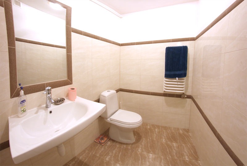 Jasna łazienka z beżowymi płytkami i brązowymi akcentami, widoczna umywalka narożna z lustrem, toaleta i grzejnik drabinkowy z niebieskim ręcznikiem.