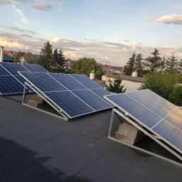 Panele fotowoltaiczne zamontowane na płaskim dachu z widokiem na zabudowę jednorodzinną i zachodzące słońce.