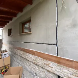 Elewacja budynku w trakcie wykończenia: położona częściowo kamienna okładzina, widoczne przewody elektryczne prowadzone po ścianie, okna z drewnianymi ramami, oraz materiały budowlane w kartonach.