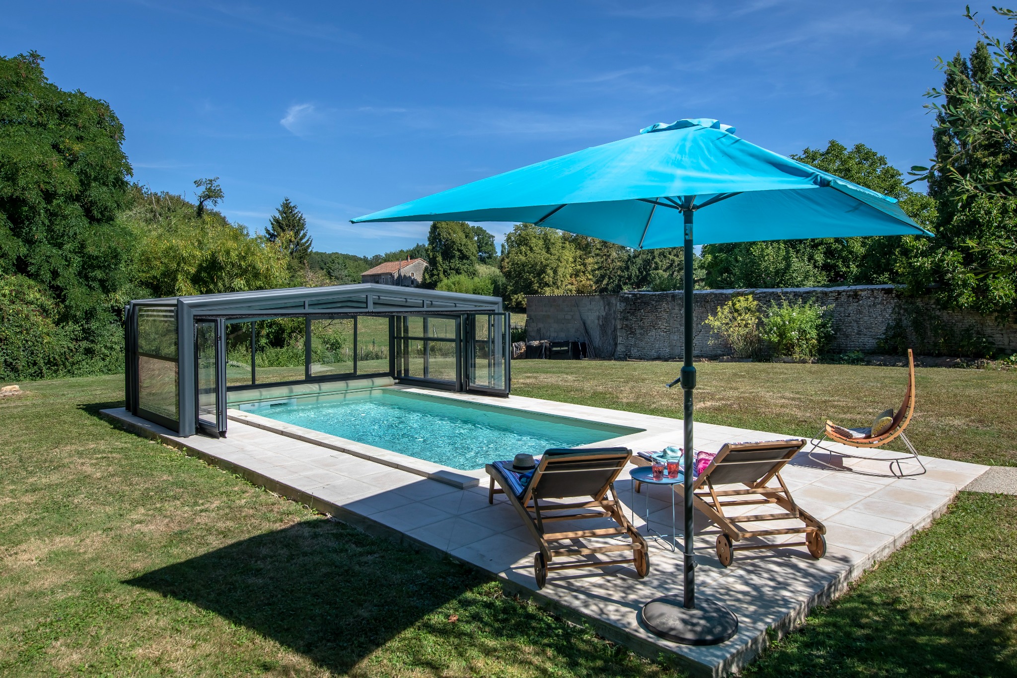 Zewnętrzny basen z przesuwną szklaną obudową, otoczony trawnikiem i kamiennym tarasem, z dwoma leżakami, stolikiem i błękitnym parasolem, w tle widoczny kamienny mur i zieleń.