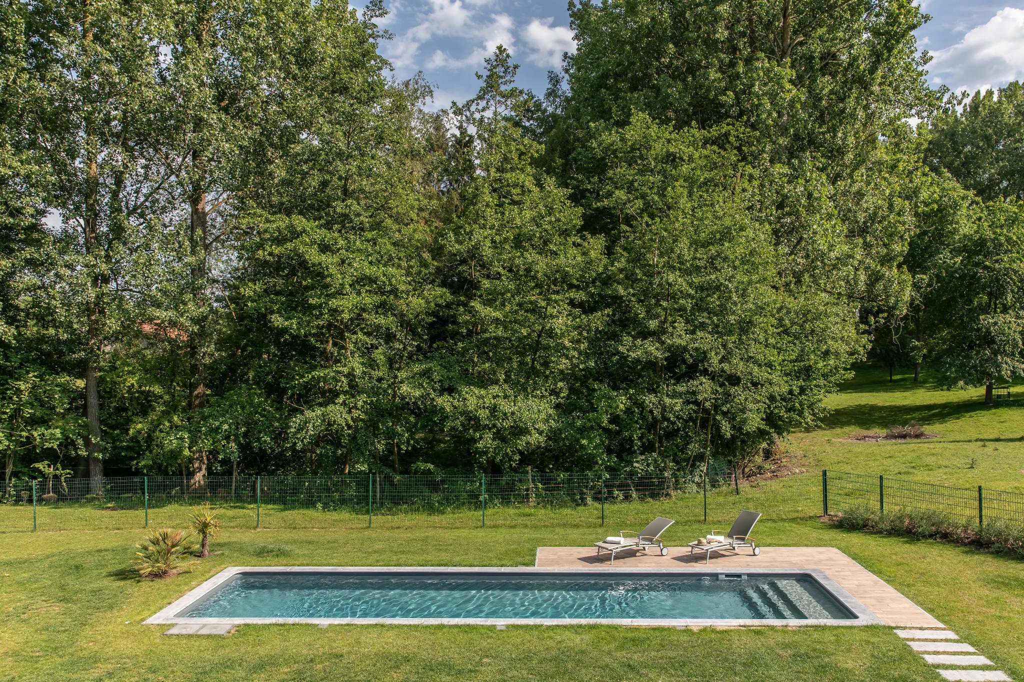 Prostokątny, nowoczesny basen w ogrodzie z zielonym trawnikiem, otoczony drzewami i zielonym ogrodzeniem z dwoma leżakami obok.