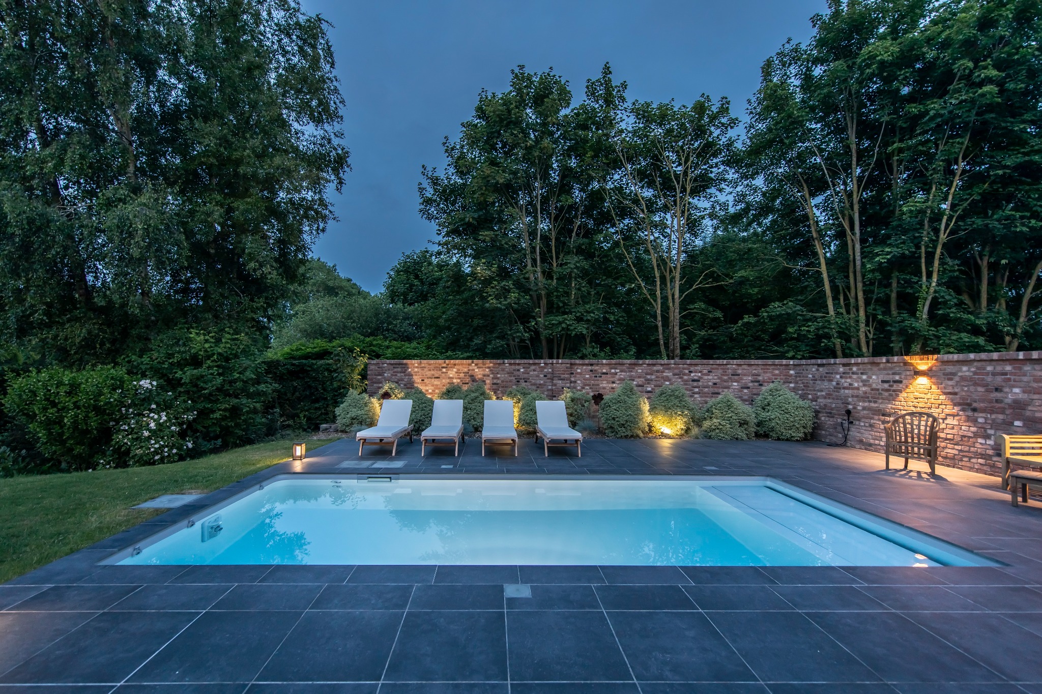 Nowoczesny basen z szarymi płytkami, czterema leżakami i oświetloną ceglaną ścianą w tle, sfotografowany o zmierzchu.