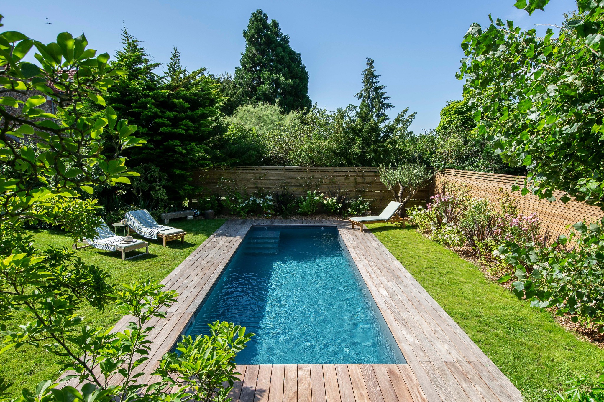 Prostokątny basen z ciemną wodą otoczony drewnianym tarasem i bujną zielenią, widok z góry, dwa leżaki z poduszkami w tle.