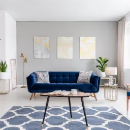 Wnętrze pokoju dziennego z granatową sofą, wzorzystym dywanem i stolikiem kawowym z otwartą książką oraz trzema abstrakcyjnymi obrazami na szarej ścianie.