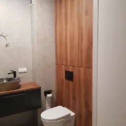 Nowoczesna łazienka z umywalką w kształcie misy na drewnianym blacie, wiszącą toaletą i wysoką szafką z drewnianymi frontami, w minimalistycznej aranżacji.