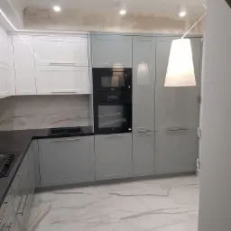 Nowoczesna kuchnia z szarymi i białymi frontami, czarnym blatem i marmurową podłogą; widoczne niedokończone prace wykończeniowe na suficie.