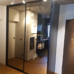 Szafa wnękowa z przesuwnymi lustrzanymi drzwiami w przedpokoju, czarna rama, widoczne odbicie kuchni, oświetlenie szynowe na suficie.