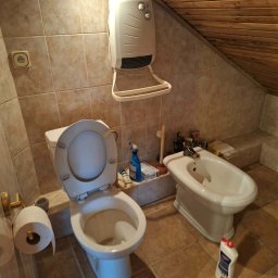 BŁYSK na PLUS - Zabrudzona toaleta i bidet w łazience z beżowymi płytkami i drewnianym sufitem. Widoczny grzejnik na ścianie i butelka środka czyszczącego na podłodze.