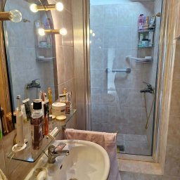 BŁYSK na PLUS - Umywalka z baterią i mydłem, szklana półka z kosmetykami, lustro z oświetleniem i kabina prysznicowa w łazience wyłożonej beżowymi płytkami. Widok na wnętrze kabiny.