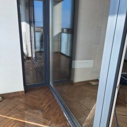 BŁYSK na PLUS - Błyszcząca, umyta szyba okna balkonowego z widokiem na zewnątrz. Na podłodze widoczne refleksy światła i ślady po myciu. W tle sprzęt do sprzątania.