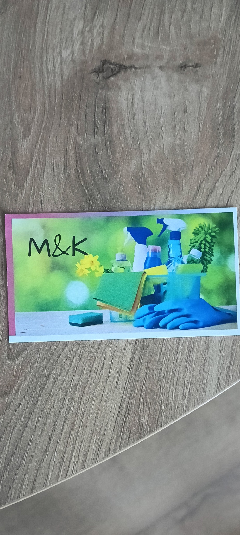 Karta z grafiką: środki czystości (płyny, gąbki, rękawice) na tle zieleni i napis 'M&K'. Karta leży na szarym, drewnianym podłożu.