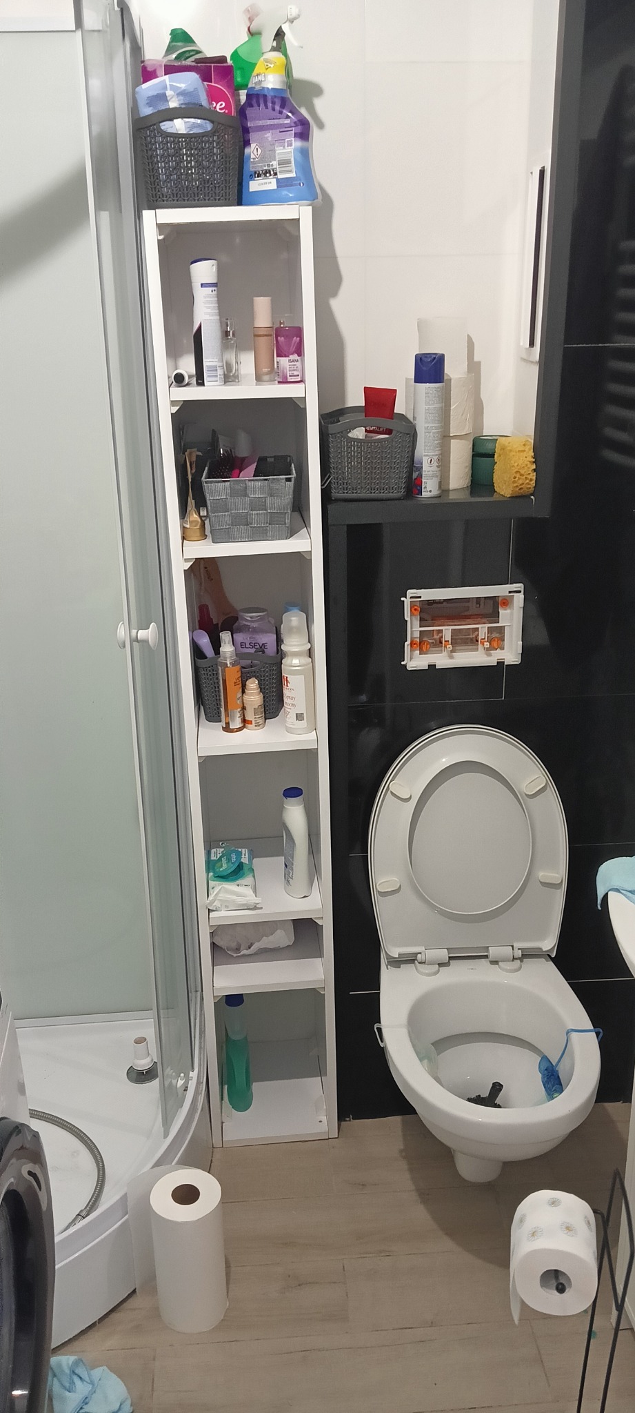 Ujęcie łazienki z otwartą toaletą, białą szafką z kosmetykami i kabiną prysznicową. Widoczne akcesoria łazienkowe i środki czystości.