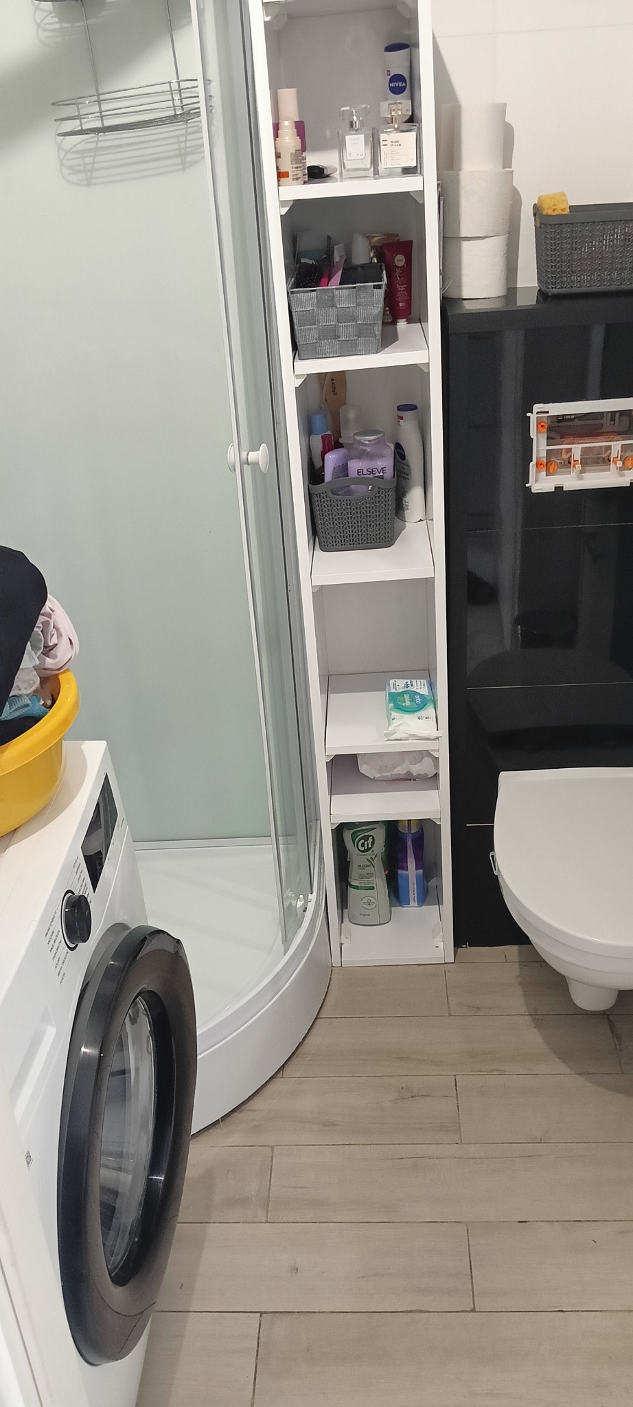 Uporządkowana łazienka z kabiną prysznicową, pralką i regałem na kosmetyki. Jasne płytki podłogowe i białe wyposażenie tworzą schludny wygląd.