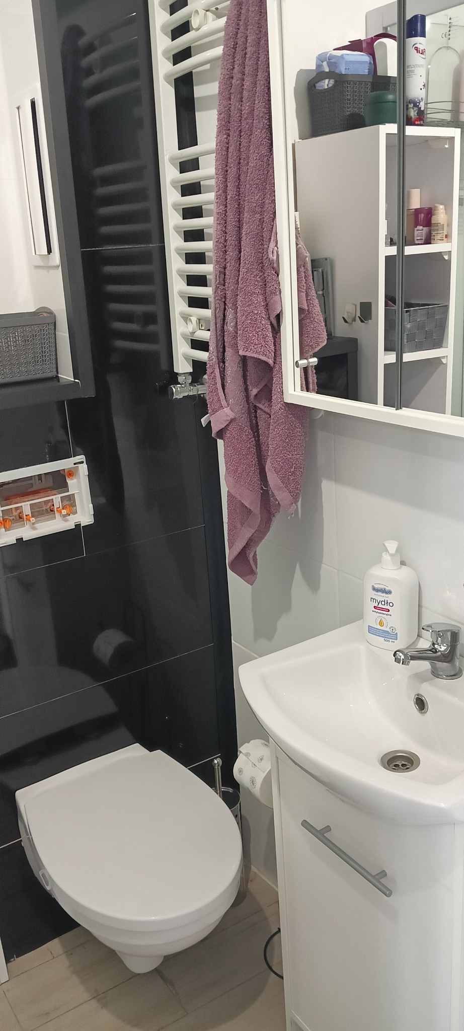Nowoczesna łazienka z czarnymi płytkami, białą ceramiką i grzejnikiem drabinkowym. Różowy ręcznik na grzejniku dodaje koloru. Widoczna umywalka z szafką i lustro.