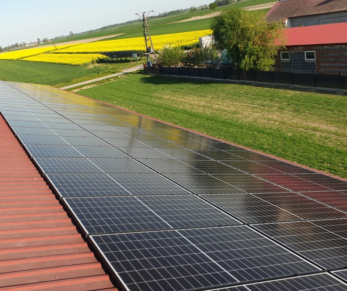 Instalacja paneli słonecznych na czerwonym dachu budynku gospodarczego, w tle pola uprawne i budynek mieszkalny.