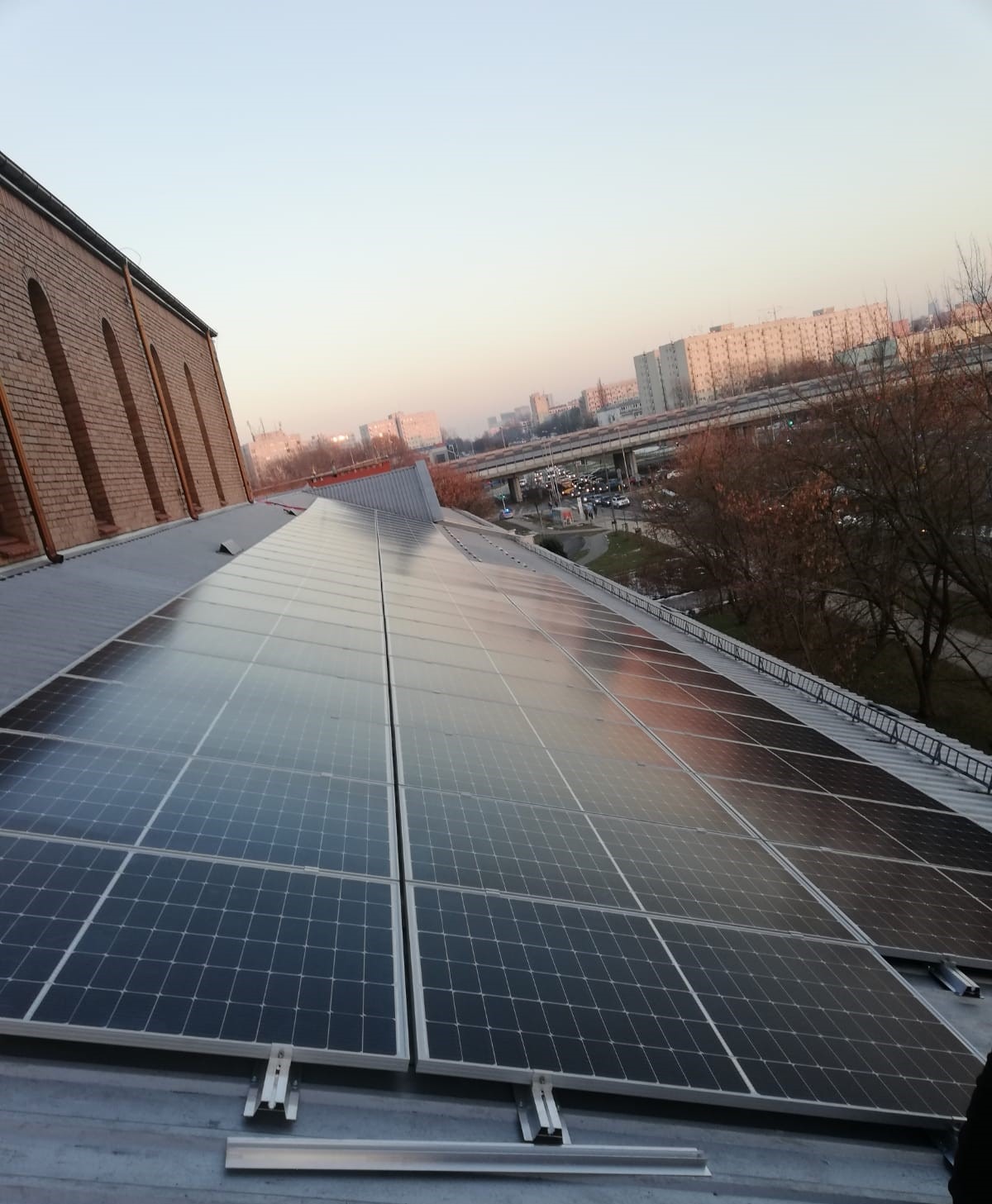 Panele słoneczne na dachu budynku z cegły, w tle widok na miasto i estakadę.